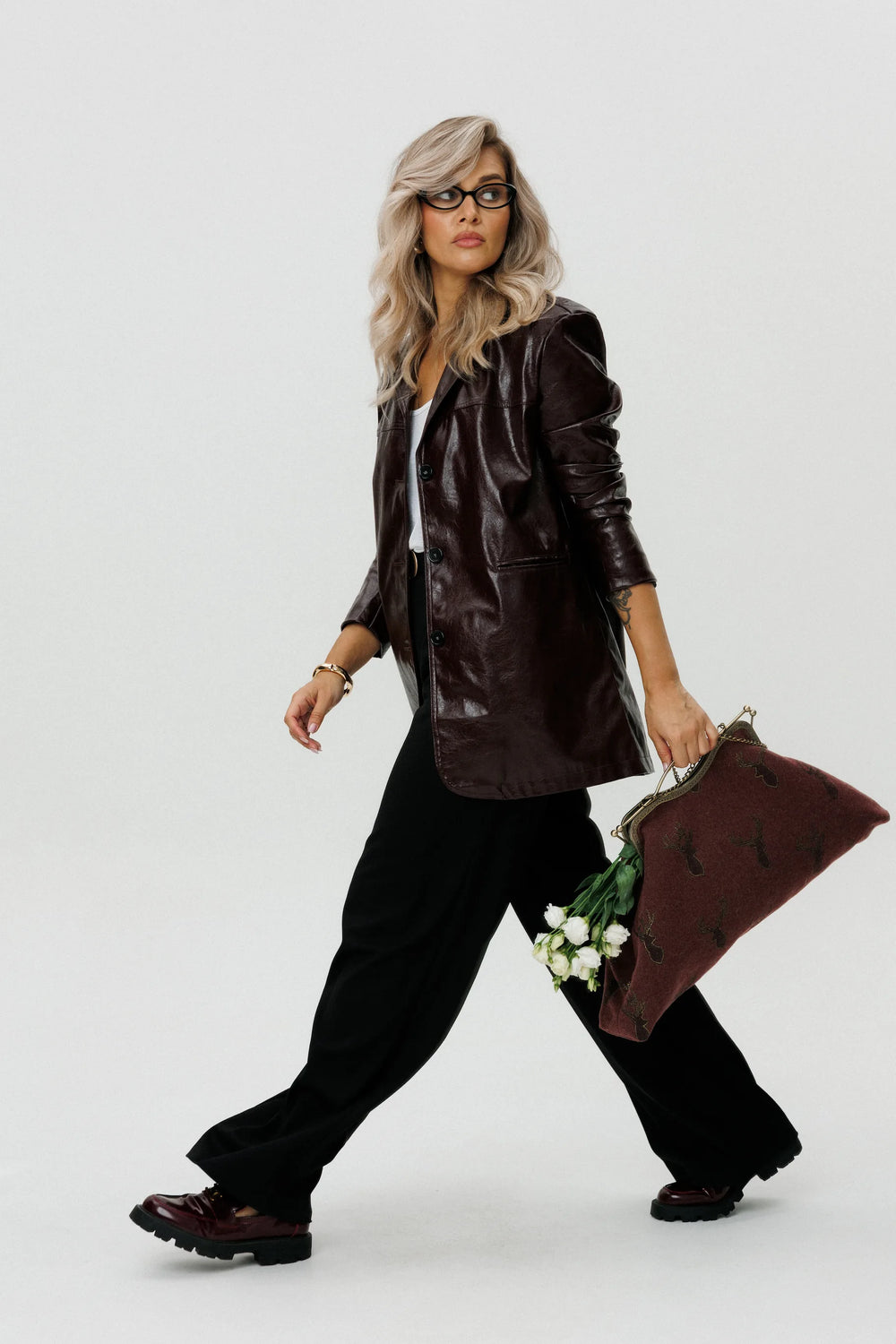Woman in a brown leather jacket and black pants holding a brown bag with white flowers on a white background