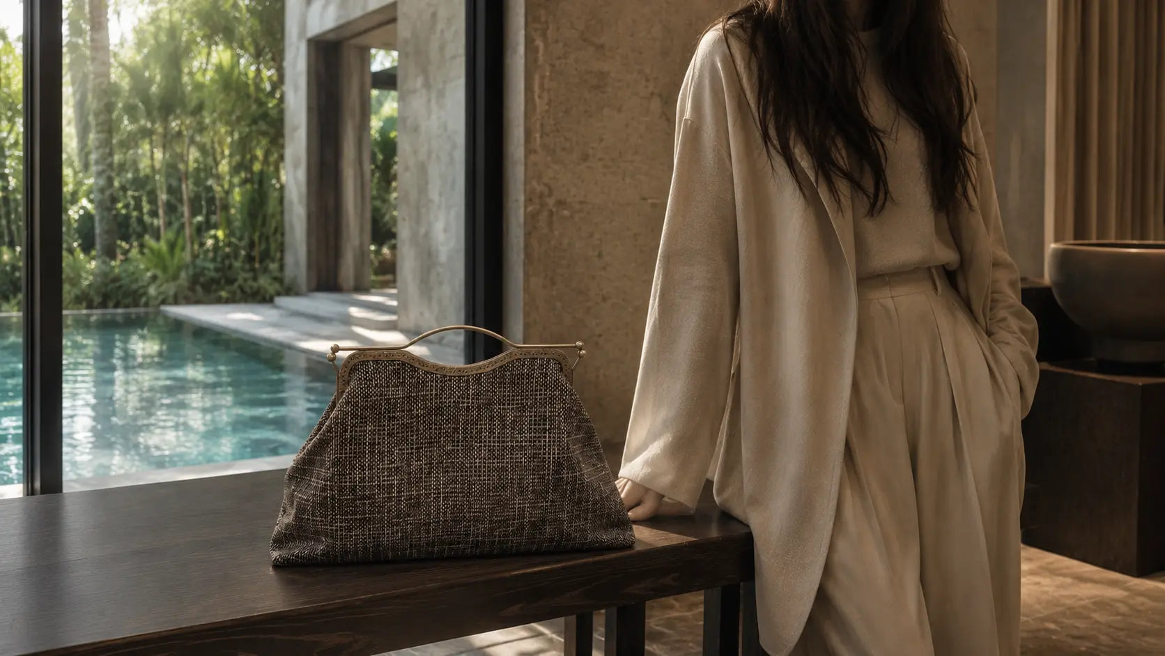 Woman in a beige outfit standing by a poolside with a textured bag on a table.